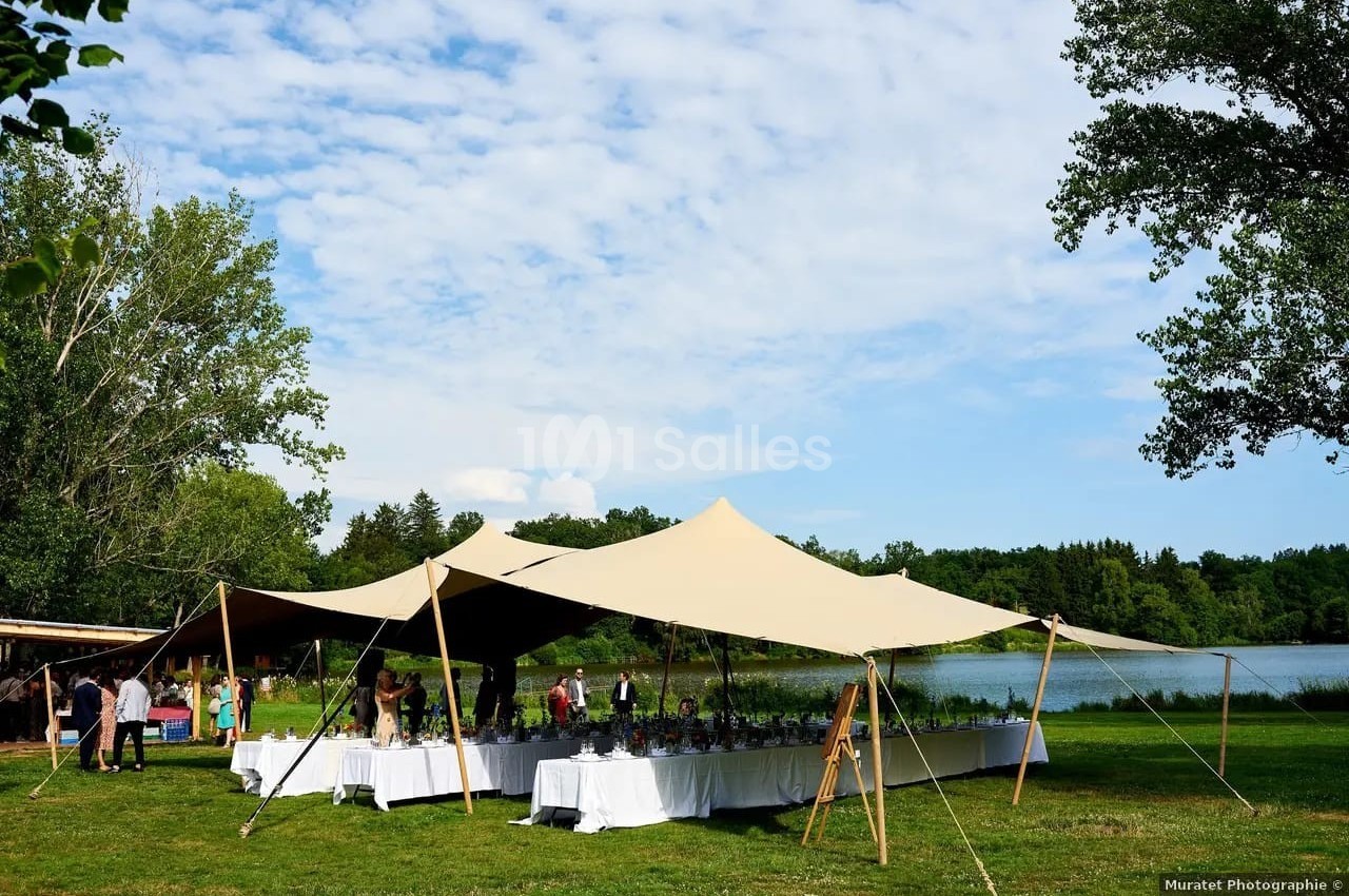 Tentes beige installées près d'un lac, avec des tables dressées pour un événement en plein air.