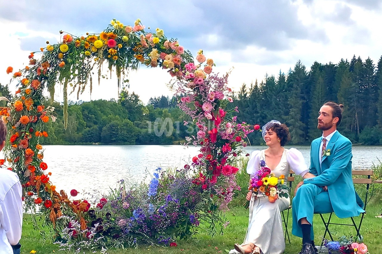 Un couple assis près d'une arche florale colorée au bord d'un lac, entouré de nature et d'un ciel nuageux.