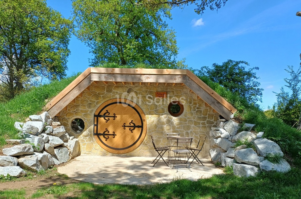 Entrée d'une maison semi-enterrée en pierre avec une porte ronde en bois, entourée de verdure et de rochers.