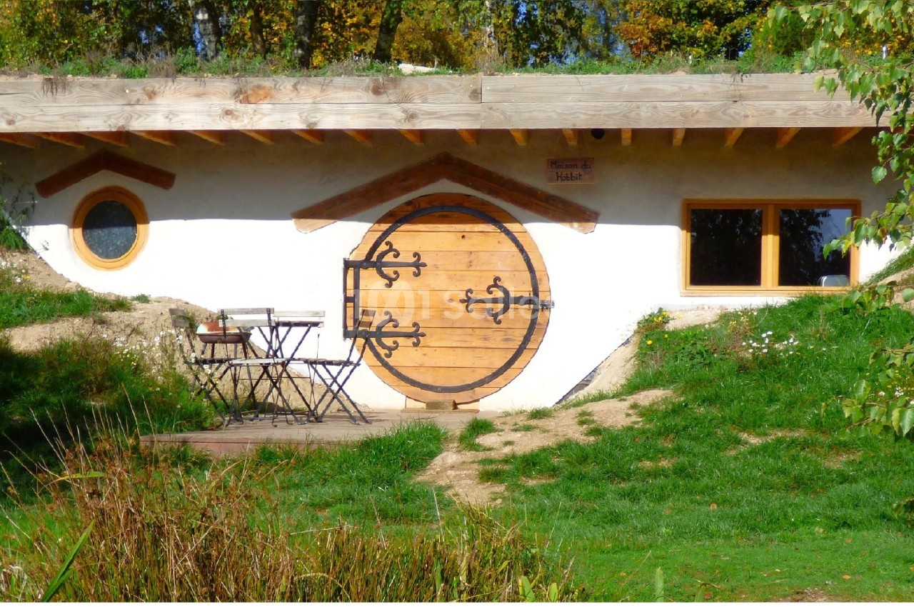 Maison semi-enterrée avec une porte ronde en bois, entourée de végétation et équipée d'une table avec deux chaises.