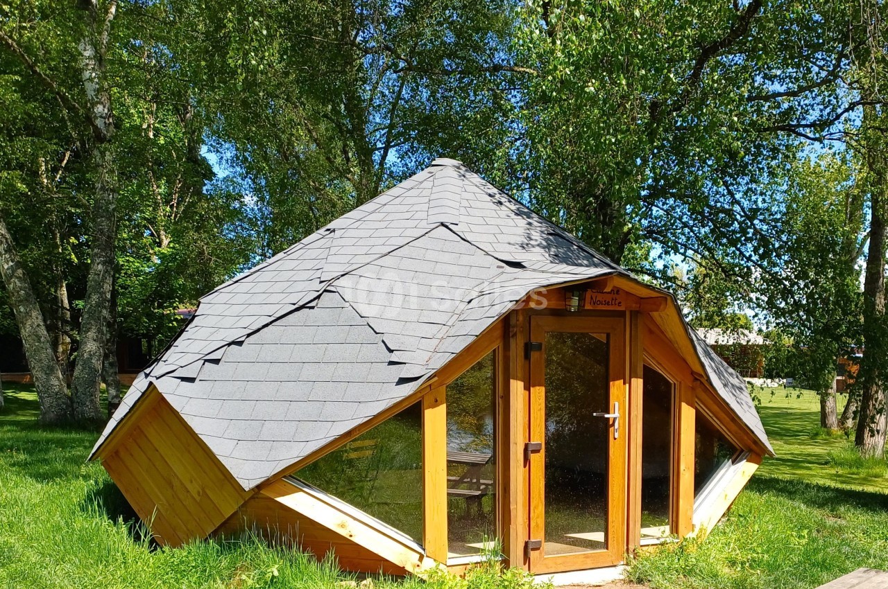 Cabane en bois au toit incliné recouvert d'ardoises, entourée d'arbres et située sur une pelouse ensoleillée.
