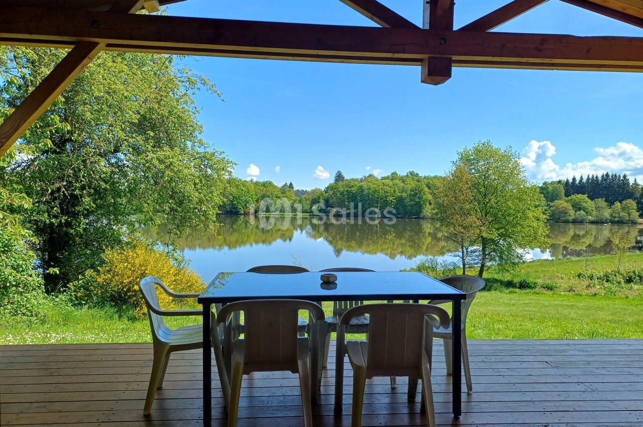 Vue sur un lac entouré de verdure depuis une terrasse en bois avec une table et des chaises en plastique.