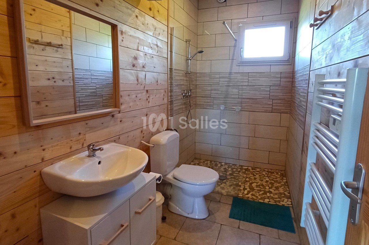 Salle de bain avec murs en bois, lavabo, toilettes, douche à l'italienne, radiateur et fenêtre apportant de la lumière…