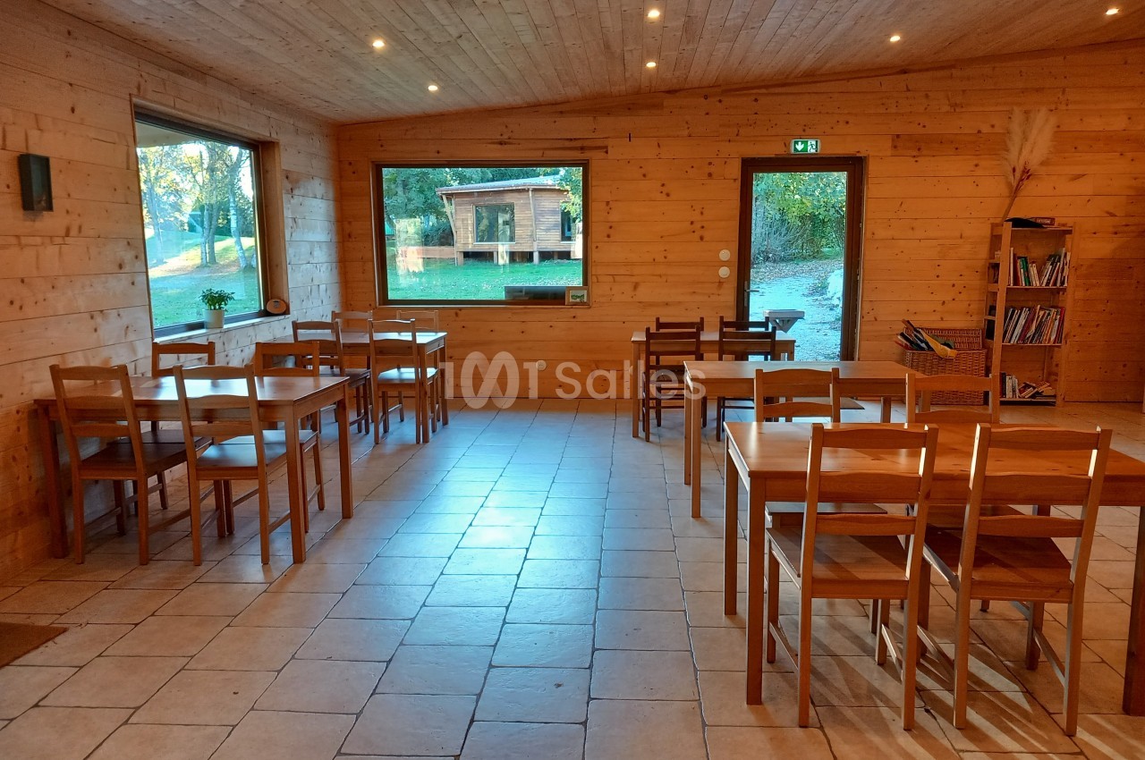 Salle en bois avec des tables et des chaises, grandes fenêtres donnant sur un extérieur verdoyant.