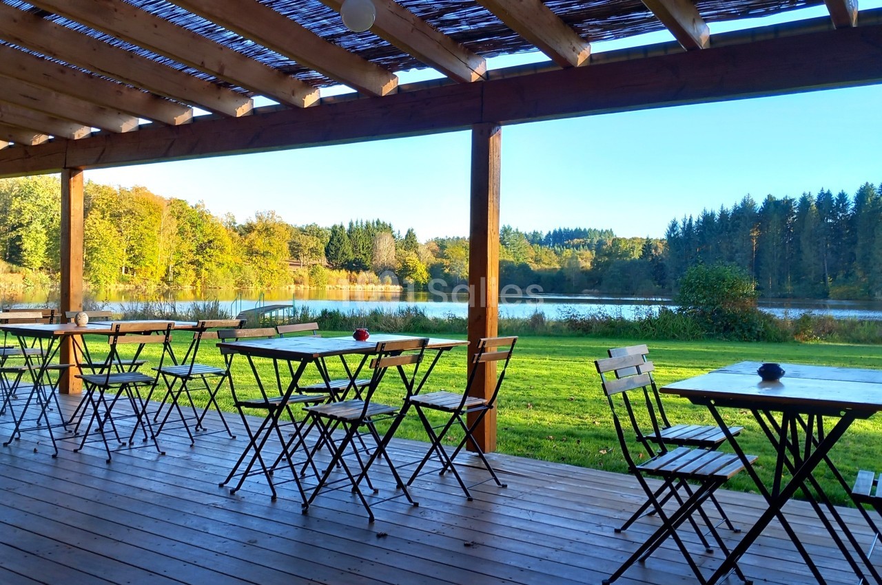Terrasse en bois avec tables et chaises donnant sur un lac entouré de verdure et d'arbres sous un ciel dégagé.