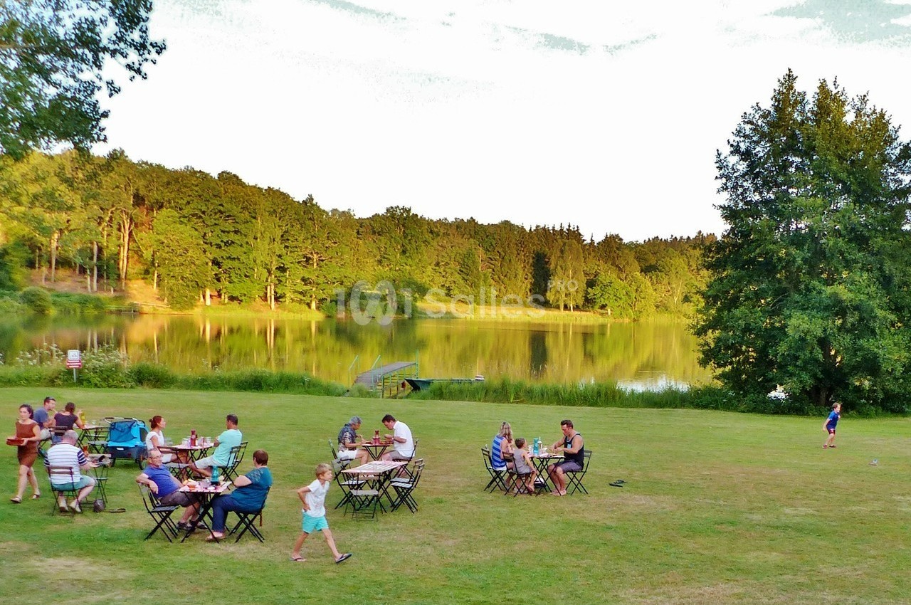 Des personnes dînent en plein air sur des tables installées près d'un lac entouré d'arbres.