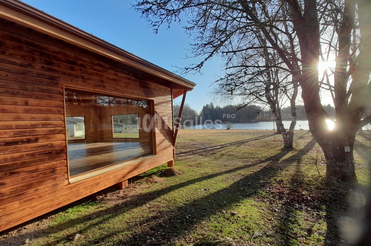 Cabane en bois avec une grande baie vitrée, située près d'un lac entouré d'arbres sous un soleil éclatant.