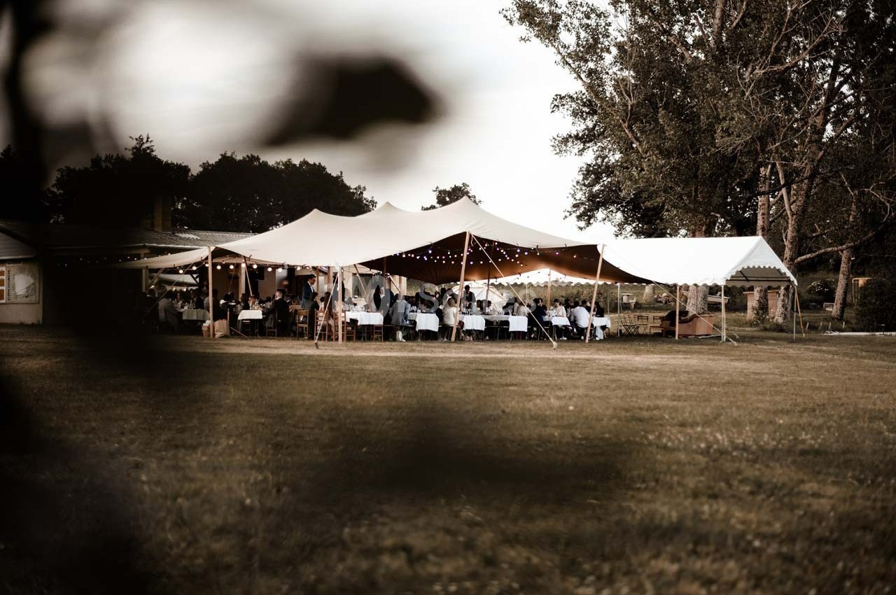 Tente blanche installée en extérieur avec des tables et des invités, entourée d'arbres et d'une pelouse.