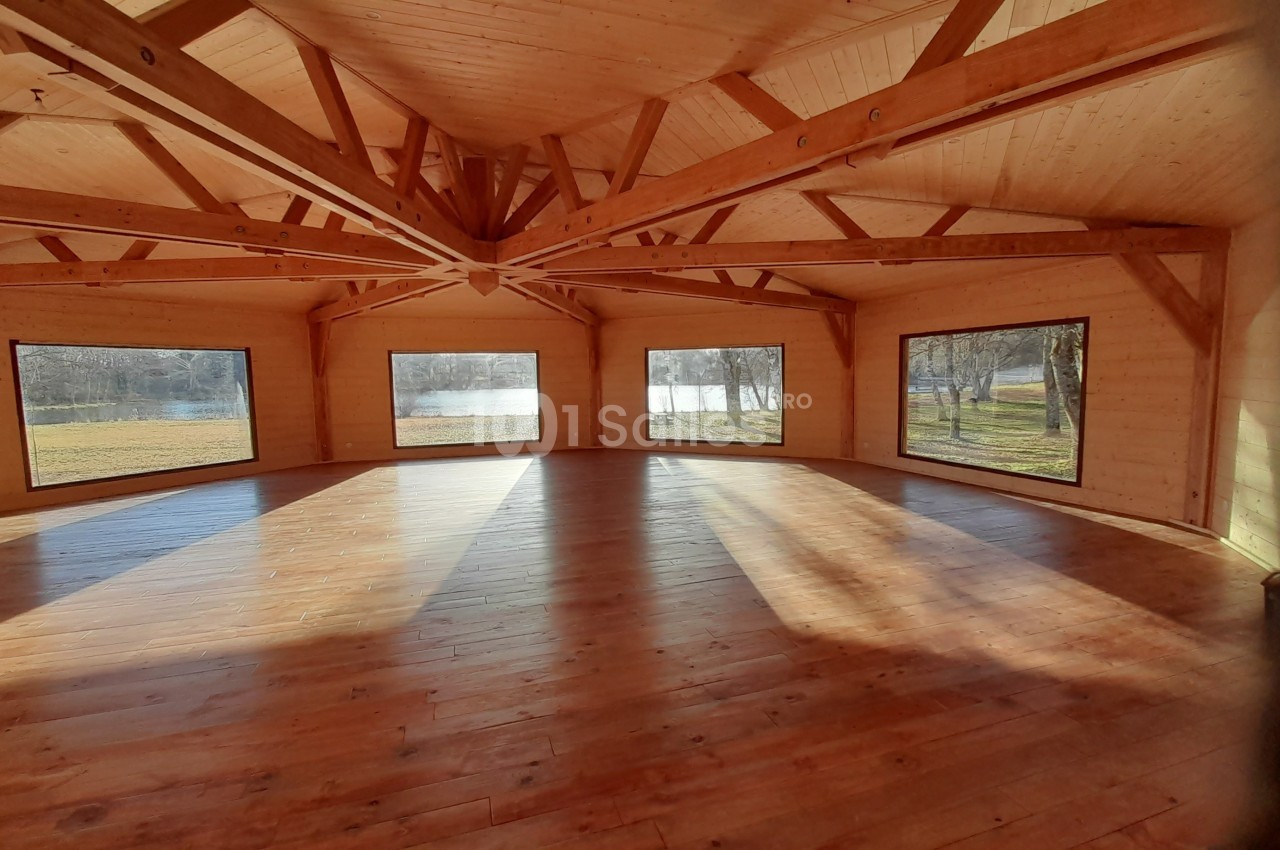 Salle en bois lumineuse avec de grandes fenêtres offrant une vue sur un paysage extérieur avec arbres et plan d'eau.