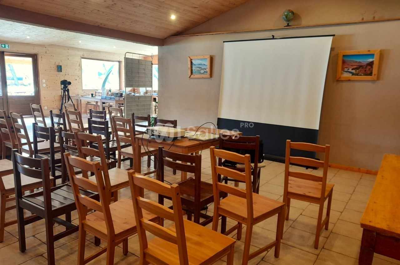 Salle avec des chaises en bois disposées face à un écran de projection dans un espace lumineux au plafond en bois.