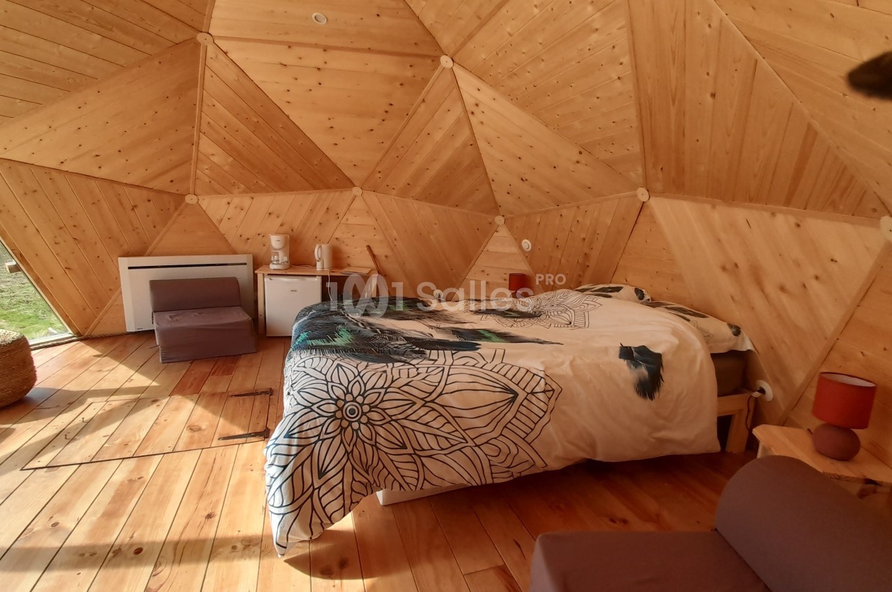 Chambre en dôme géodésique avec murs en bois, lit double, fauteuils, petite table et vue sur l'extérieur verdoyant.
