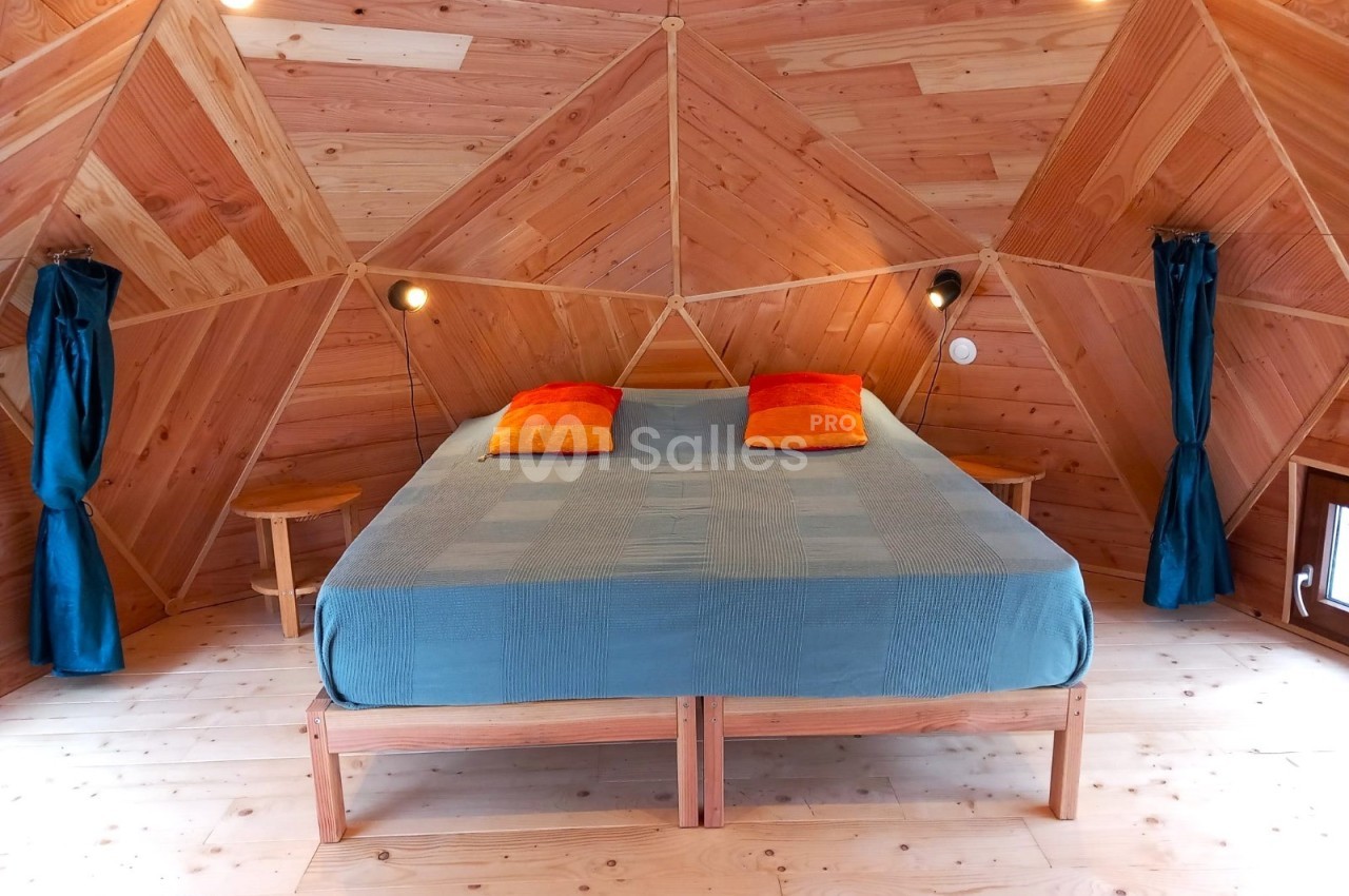 Chambre en bois avec un lit double, drap bleu, coussins orange et rideaux bleus, éclairée par des lampes murales.