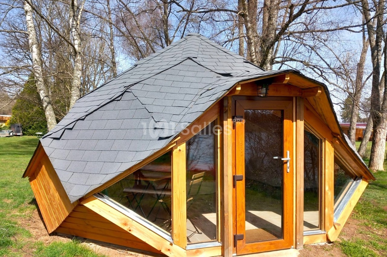 Cabane en bois avec toit en tuiles grises, grandes fenêtres et porte vitrée, située dans un espace arboré.