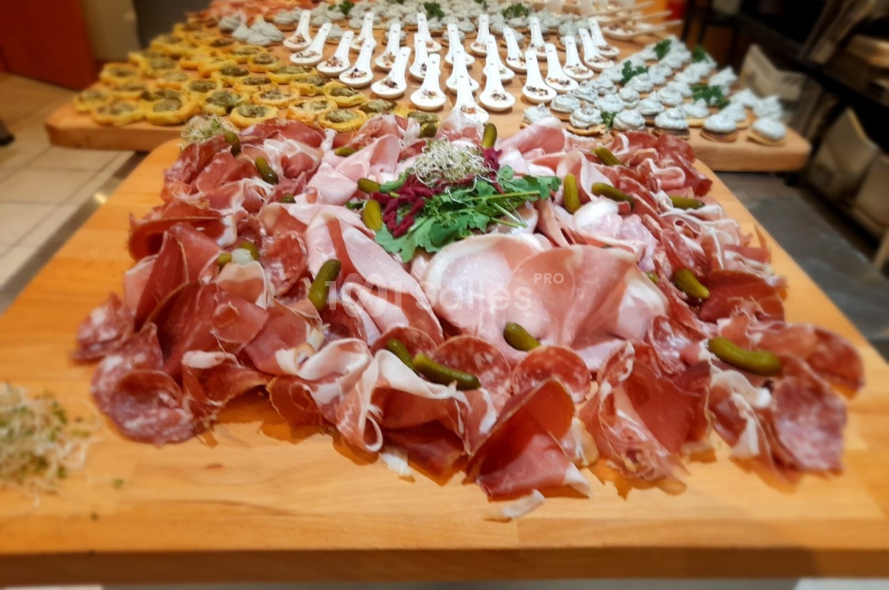 Plateau de charcuterie avec jambon, saucisson, cornichons et salade, entouré d'amuse-bouches variés en arrière-plan.