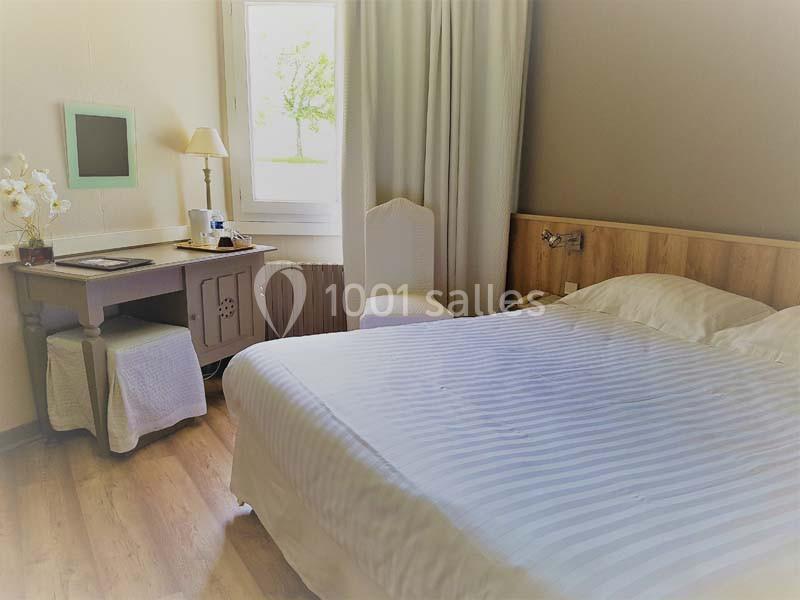 Chambre lumineuse avec lit double, bureau équipé, chaise, lampe et fenêtre donnant sur un paysage verdoyant.