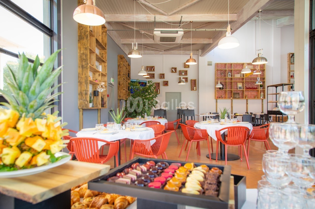 Salle de restaurant lumineuse avec tables dressées, chaises rouges et buffet de pâtisseries et fruits au premier plan.