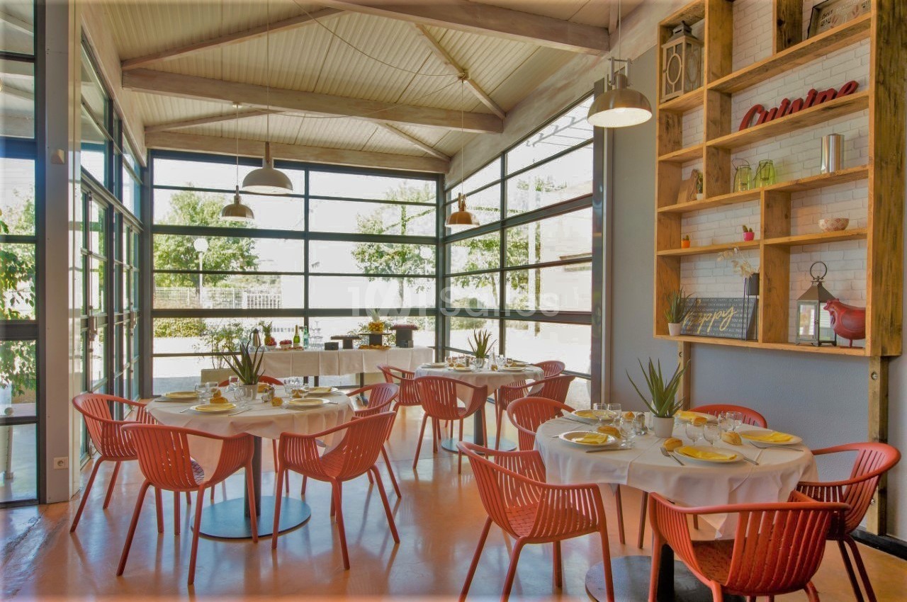 Salle lumineuse avec de grandes baies vitrées, tables dressées avec chaises rouges et étagères décoratives en bois.