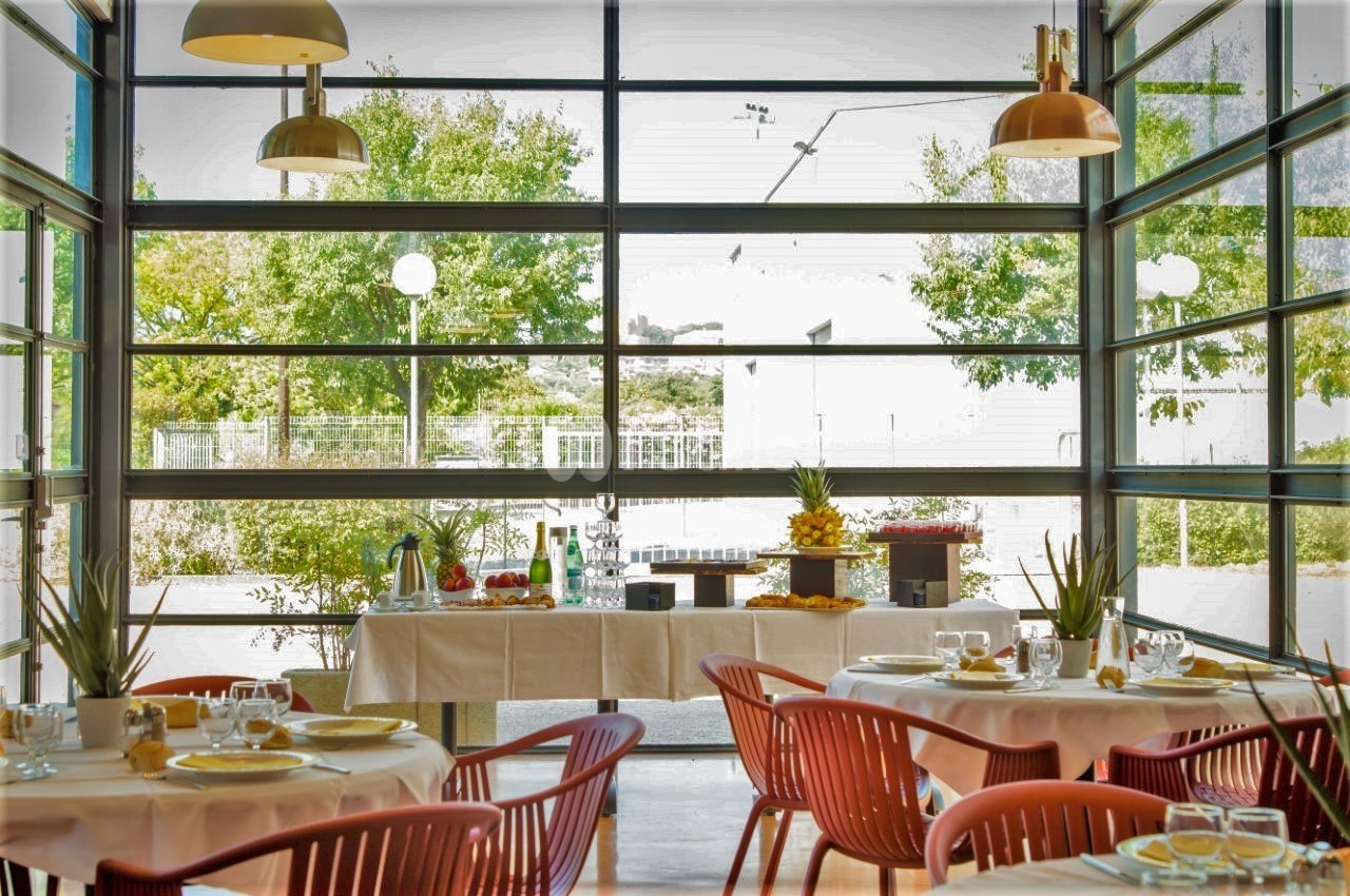 Salle de restaurant lumineuse avec grandes baies vitrées, tables dressées et buffet garni au fond.