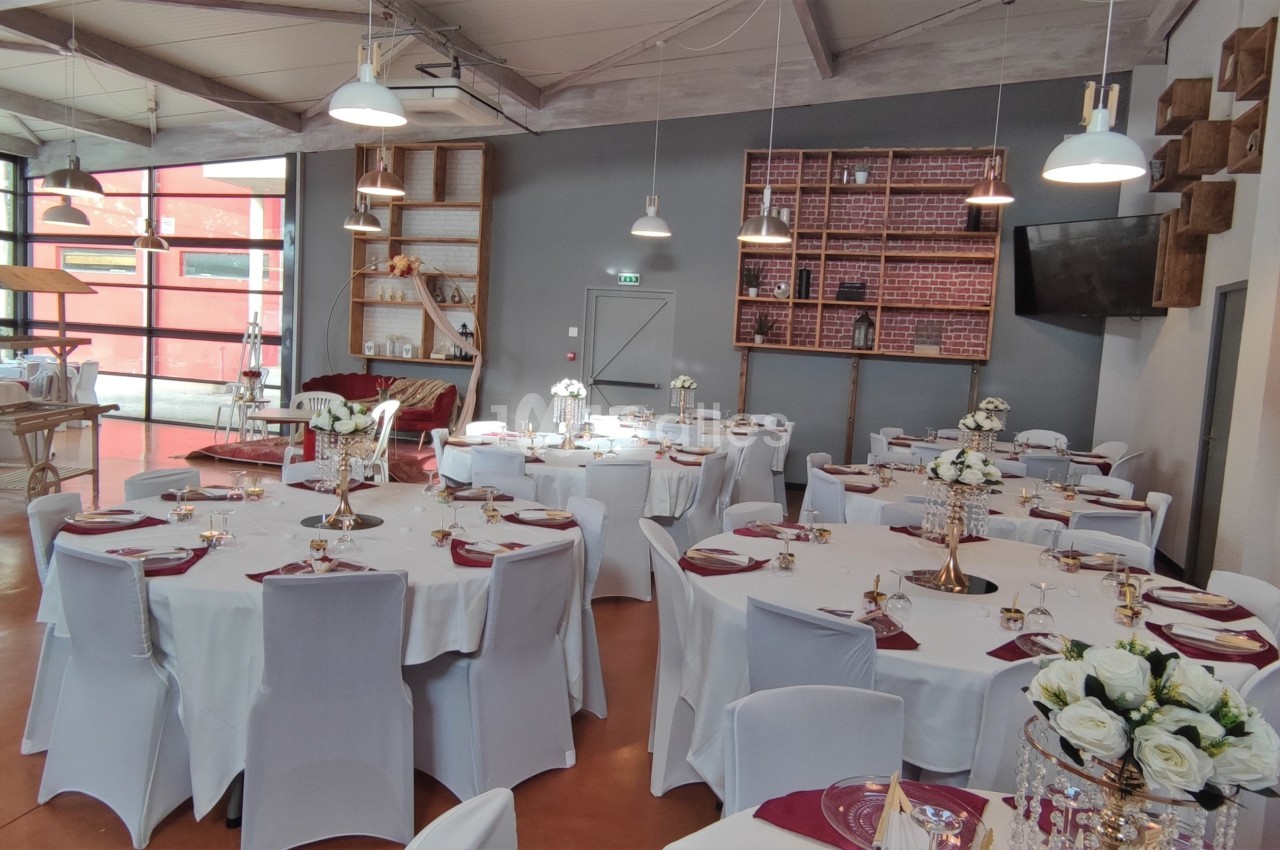 Salle de réception lumineuse avec tables rondes dressées, nappes blanches, décorations florales et étagères en arrière-plan.