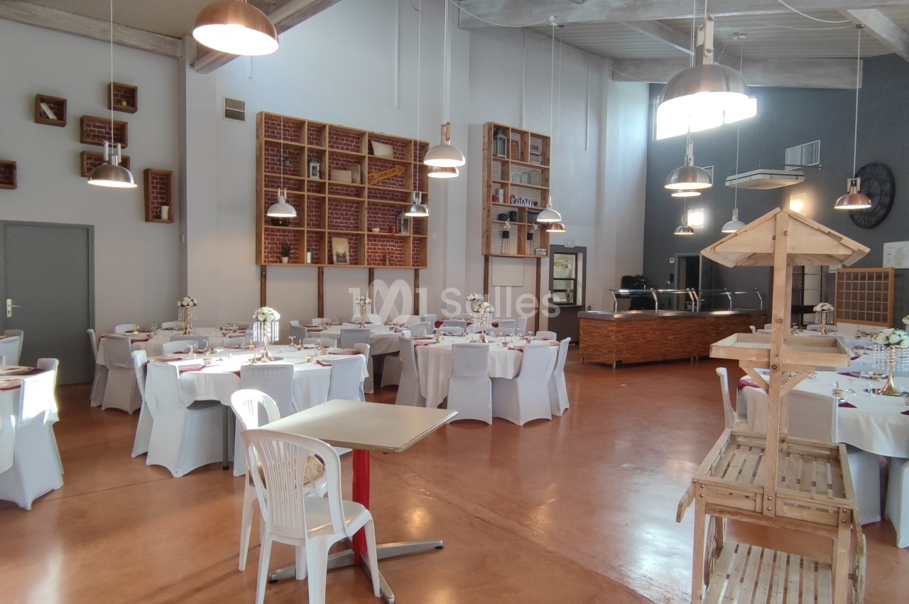 Salle de réception lumineuse avec tables rondes dressées, chaises blanches et décorations florales.