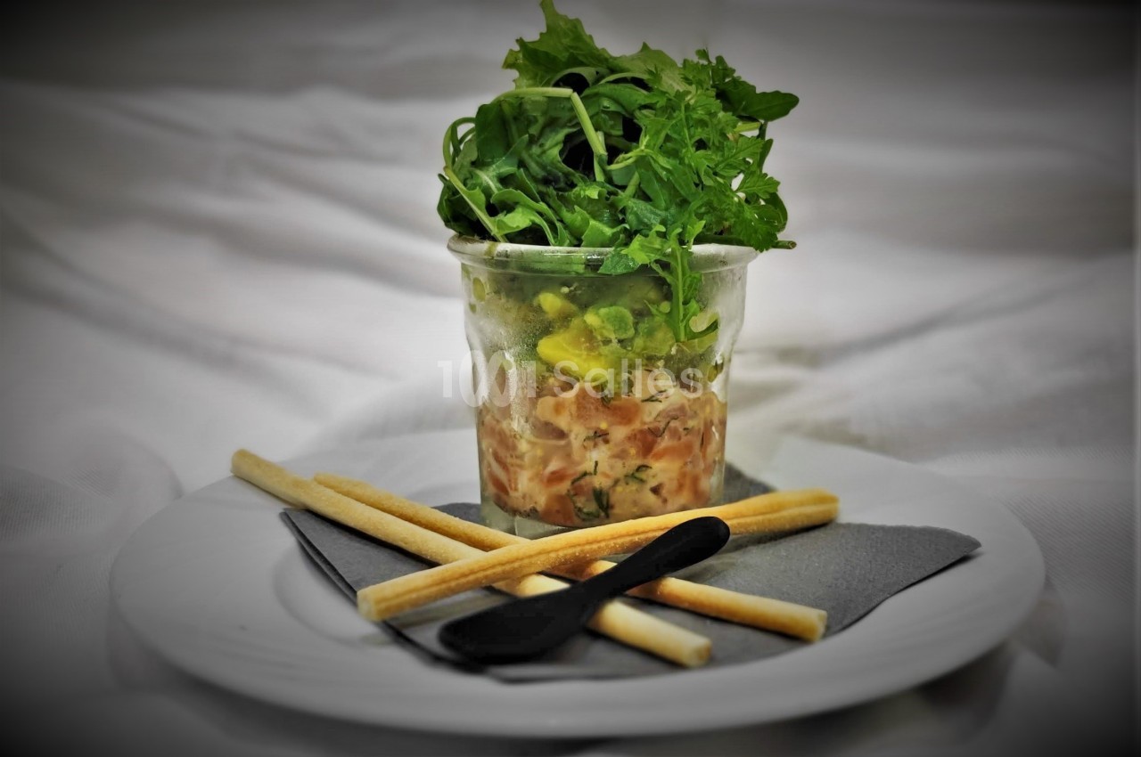 Verrine de tartare de saumon surmontée de roquette, accompagnée de gressins et d'une cuillère noire.