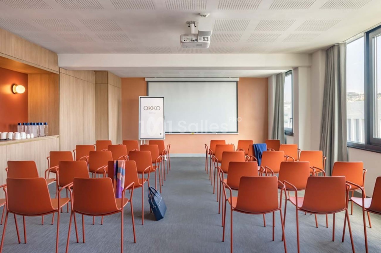 Salle de réunion lumineuse avec chaises orange, écran de projection, tableau blanc et fenêtres donnant sur l'extérieur.