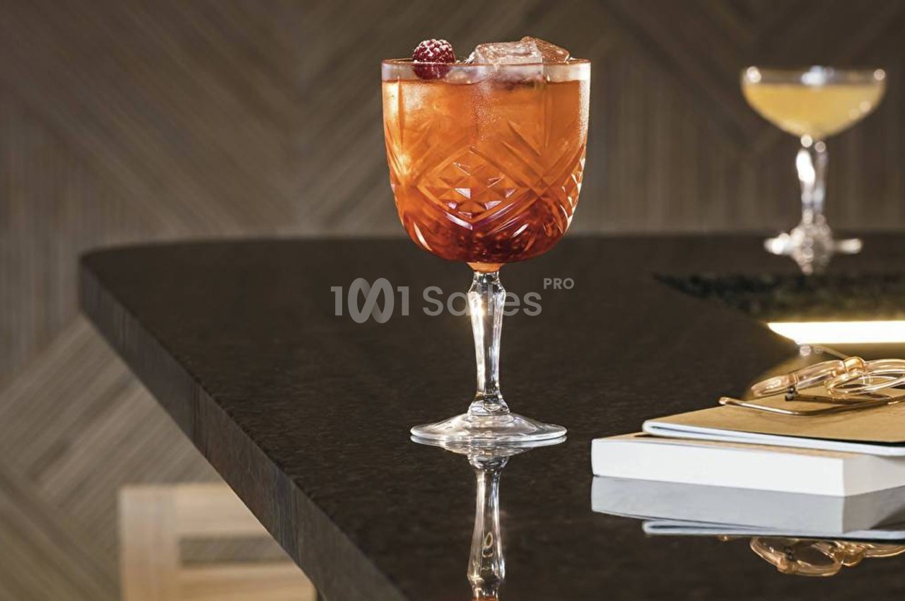 Verre à cocktail rouge garni de fruits rouges et glaçons, posé sur un comptoir sombre avec des carnets à proximité.