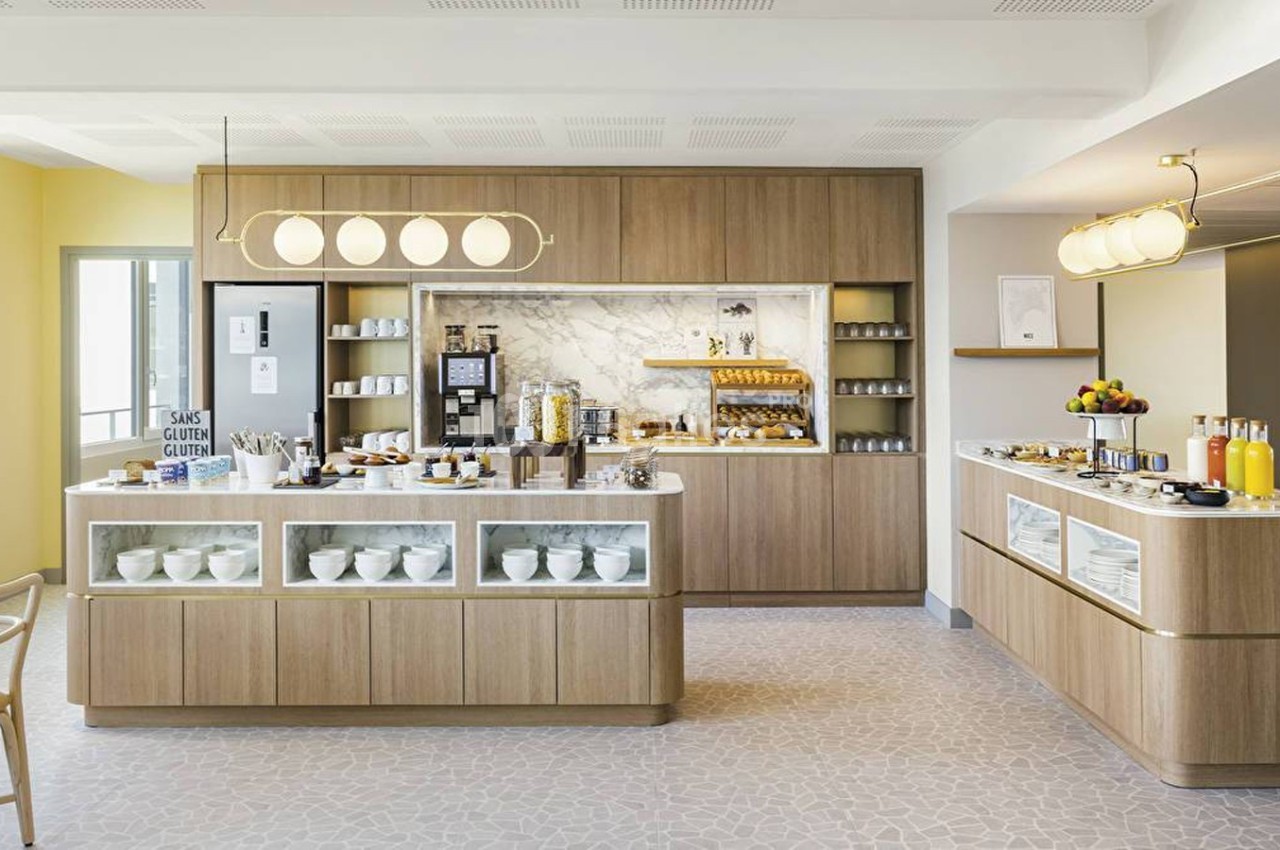 Buffet de petit-déjeuner dans une salle lumineuse avec comptoirs en bois, boissons, viennoiseries et vaisselle.
