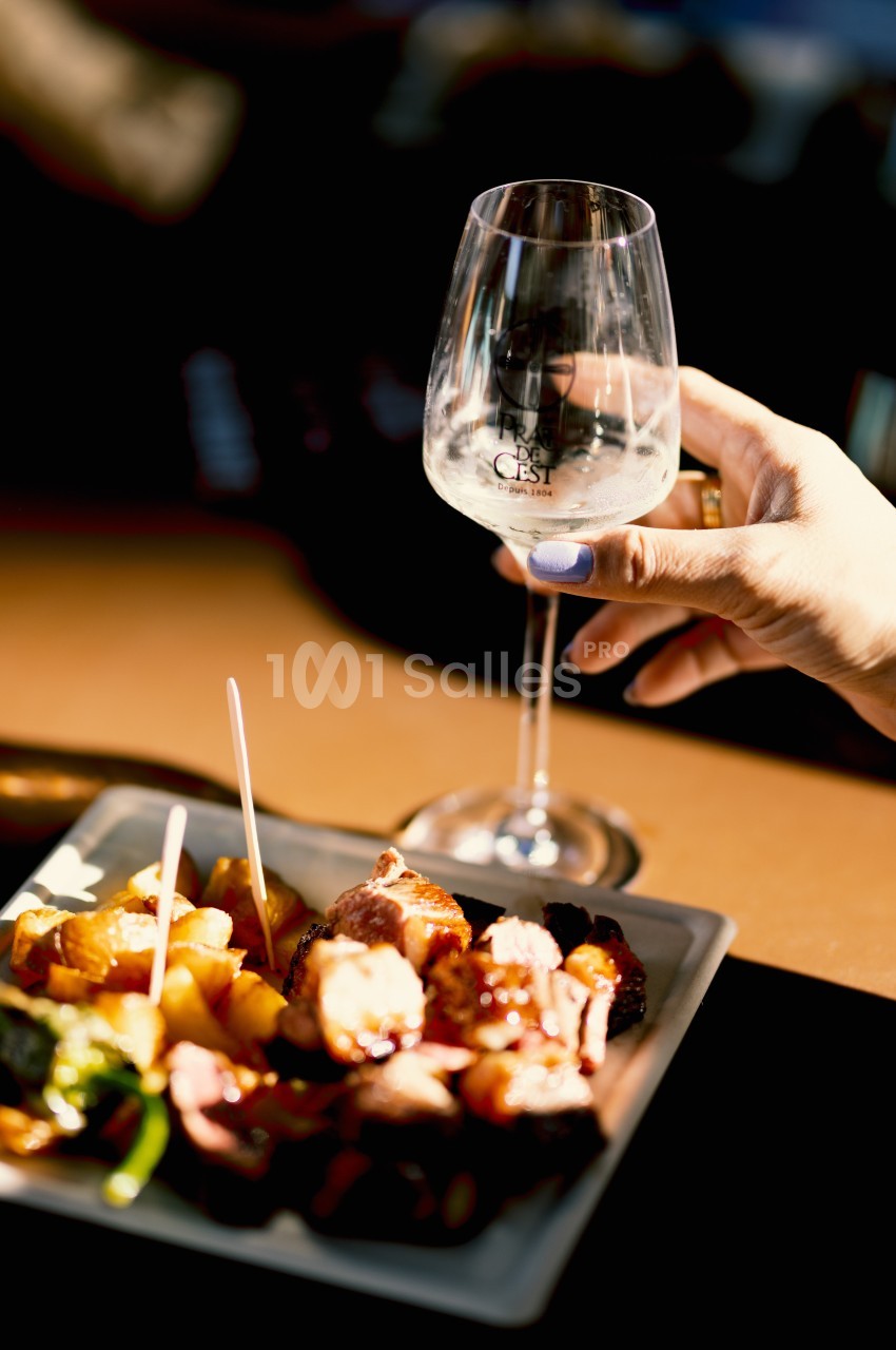 Une main tient un verre de vin blanc près d'une assiette de brochettes et légumes grillés sur une table.