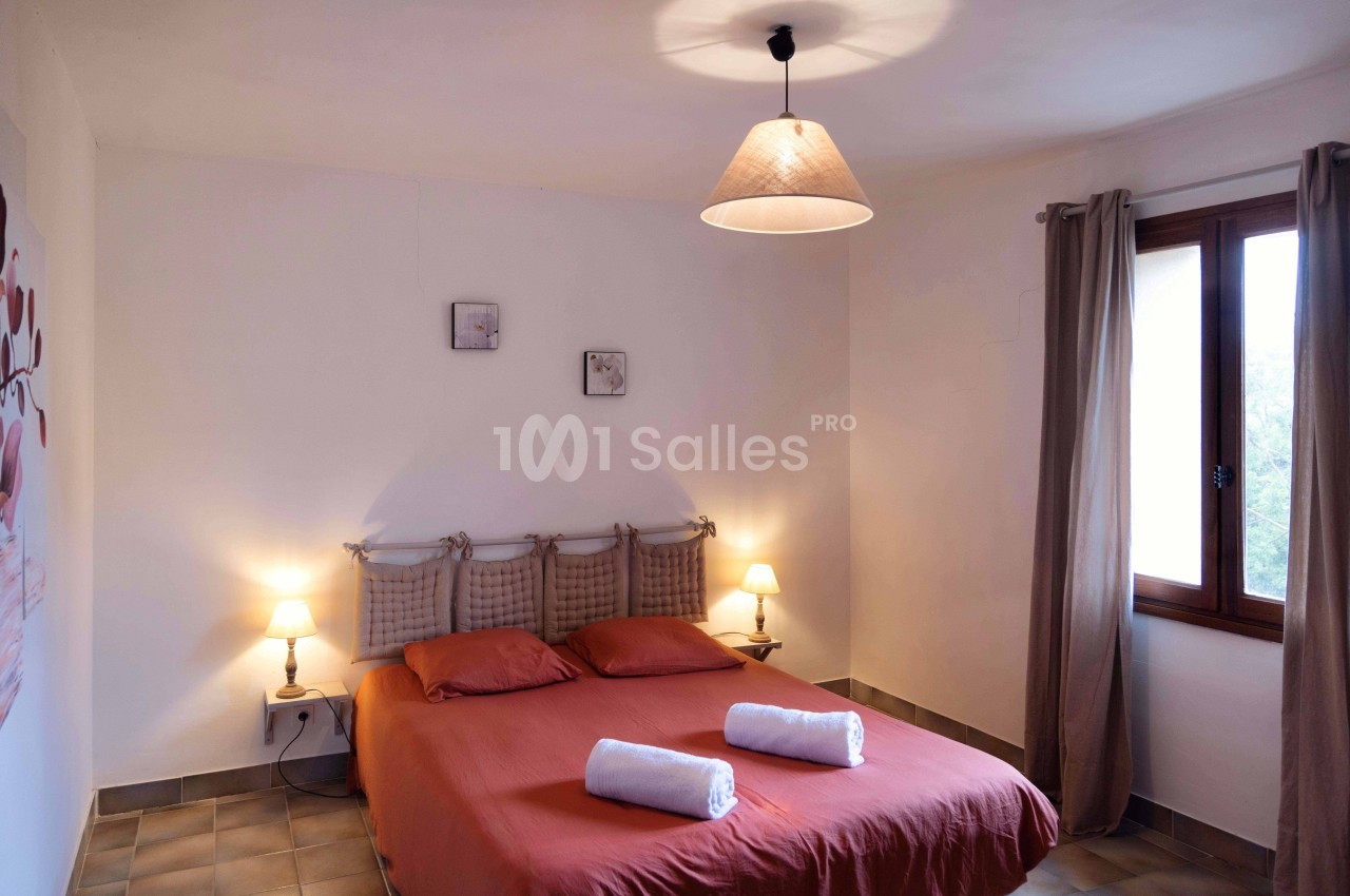 Chambre lumineuse avec lit double, draps rouges, deux serviettes pliées, lampes de chevet et fenêtre avec rideaux beiges.