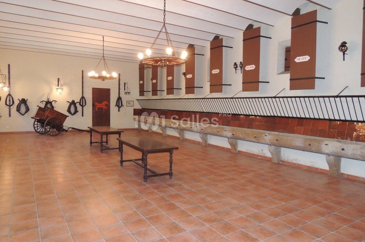 Salle avec mangeoires en pierre, tables en bois, murs décorés d'outils équestres et plafonniers suspendus.