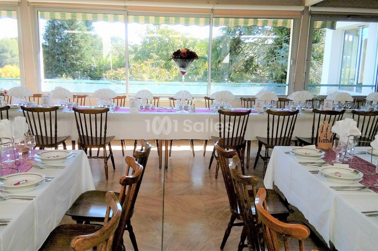 Salle de réception lumineuse avec tables dressées, nappes blanches, chaises en bois et vue sur un jardin à travers de…