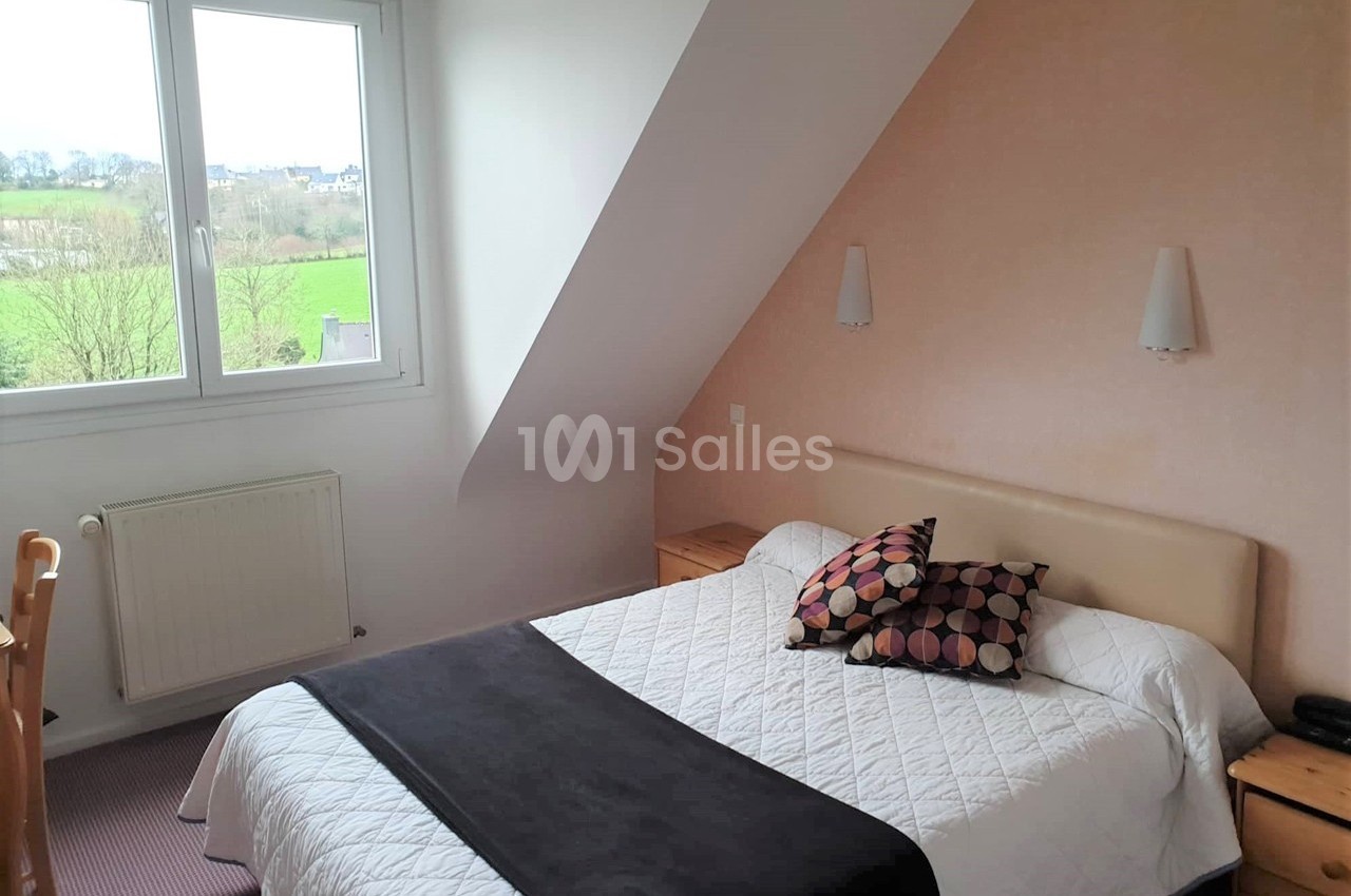 Chambre lumineuse avec lit double, murs clairs, bureau en bois, et vue sur un paysage verdoyant par la fenêtre.