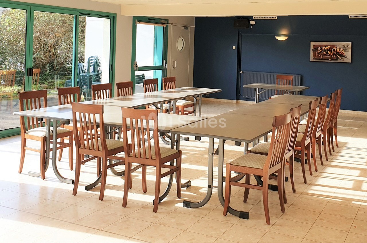 Salle lumineuse avec tables rectangulaires et chaises en bois disposées en U, grandes baies vitrées sur la gauche.