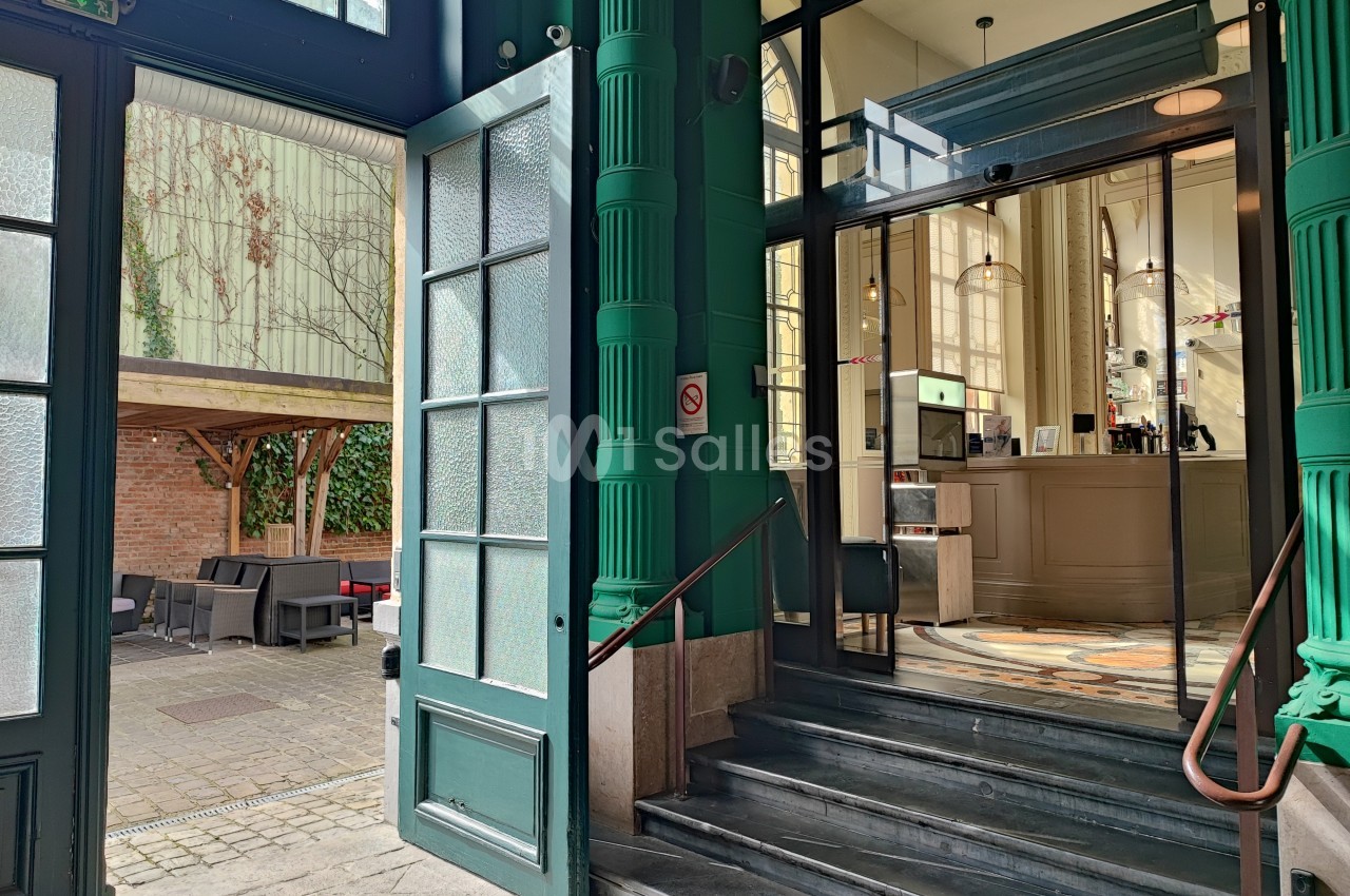 Entrée d'un bâtiment avec des portes ouvertes, escalier menant à un intérieur lumineux et terrasse extérieure aménagée.