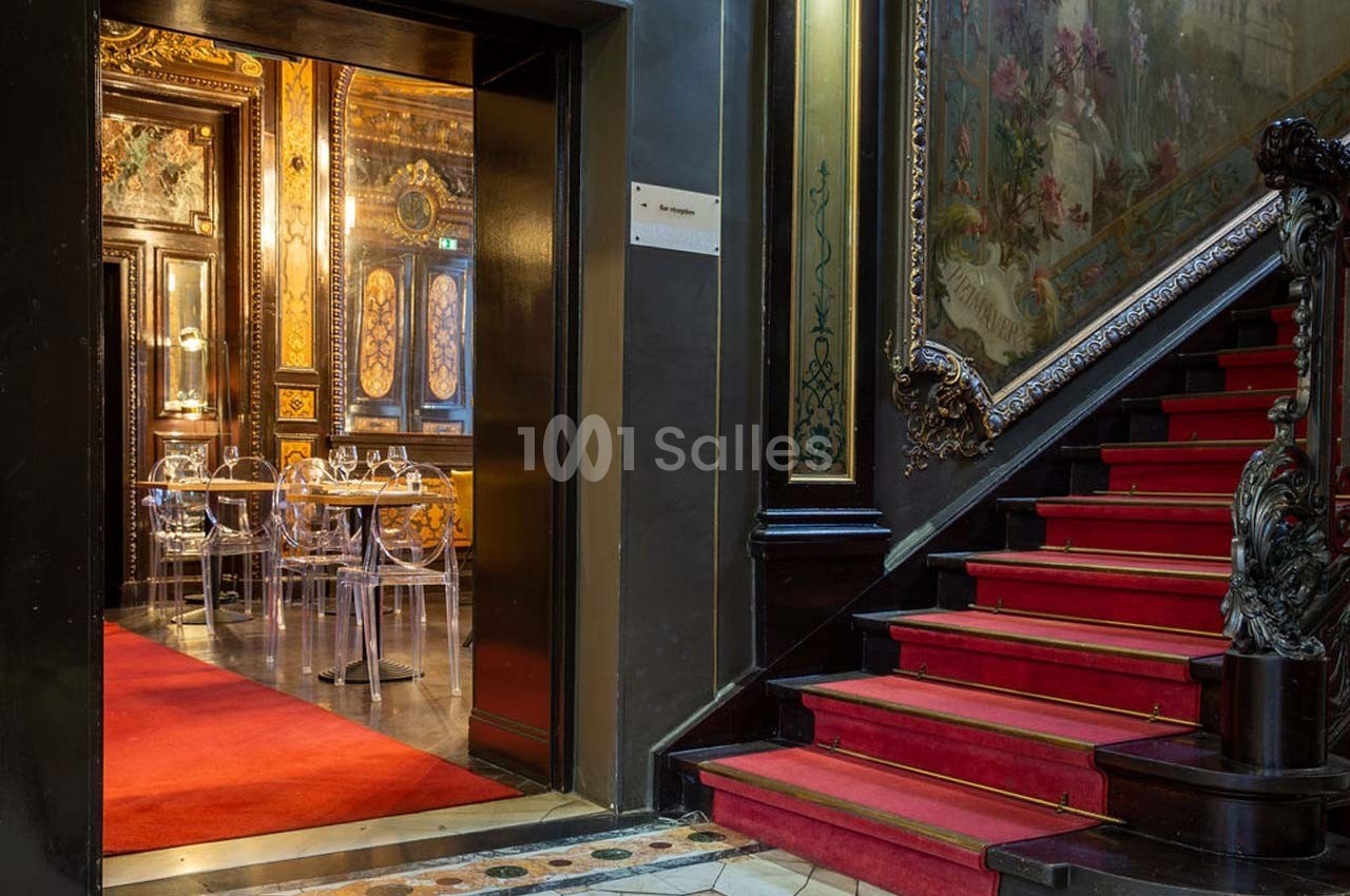 Escalier en tapis rouge menant à une salle ornée de décorations murales et de chaises transparentes.