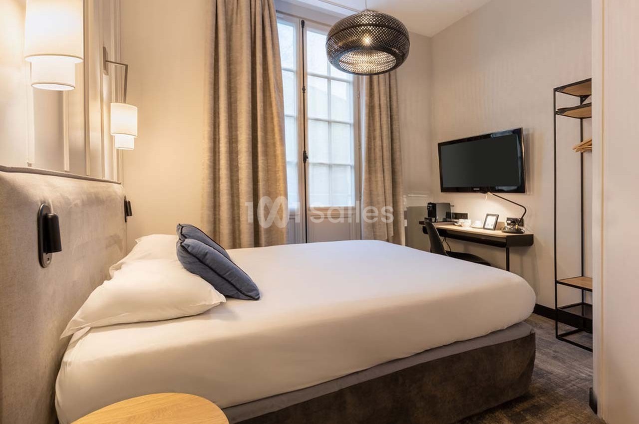 Chambre lumineuse avec lit double, bureau, télévision murale, lampes modernes et rideaux devant une fenêtre.