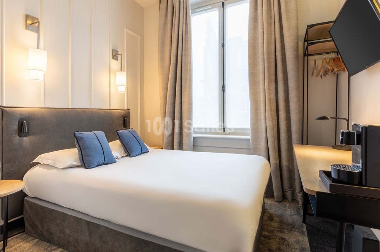 Chambre d'hôtel lumineuse avec un lit double, des coussins bleus, un bureau, une télévision et une grande fenêtre.