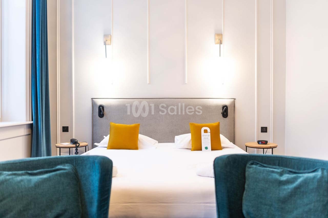 Lit double avec tête de lit grise, coussins jaunes, lampes murales et fauteuils bleus dans une chambre lumineuse.