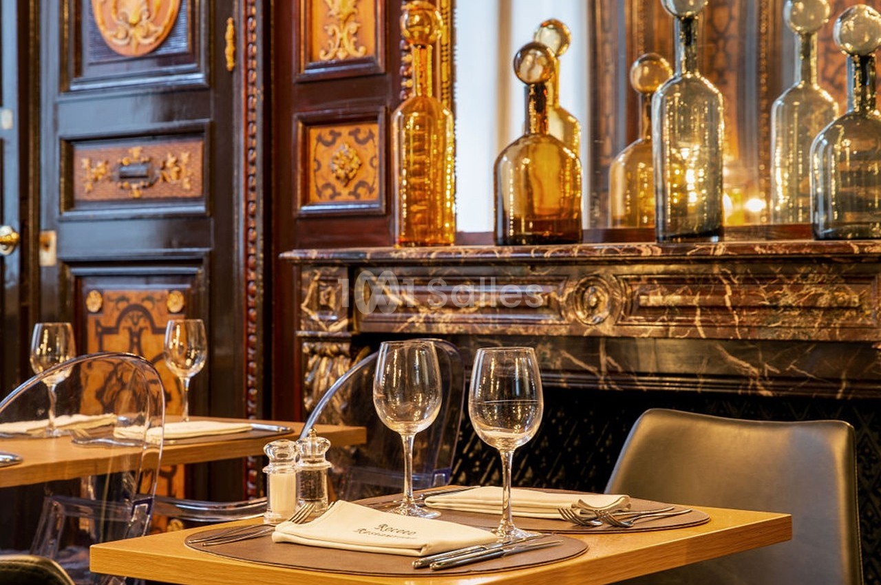 Tables dressées avec verres et couverts dans un restaurant élégant, décoré de boiseries et de bouteilles en verre.