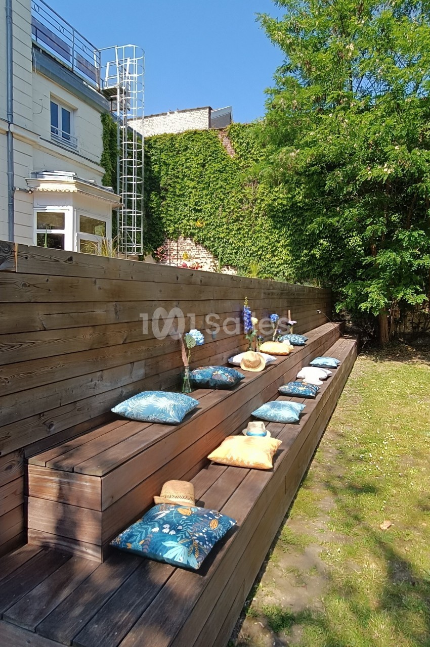 Bancs en bois avec coussins colorés disposés en terrasse dans un jardin ensoleillé, bordé d'arbres et d'une maison.