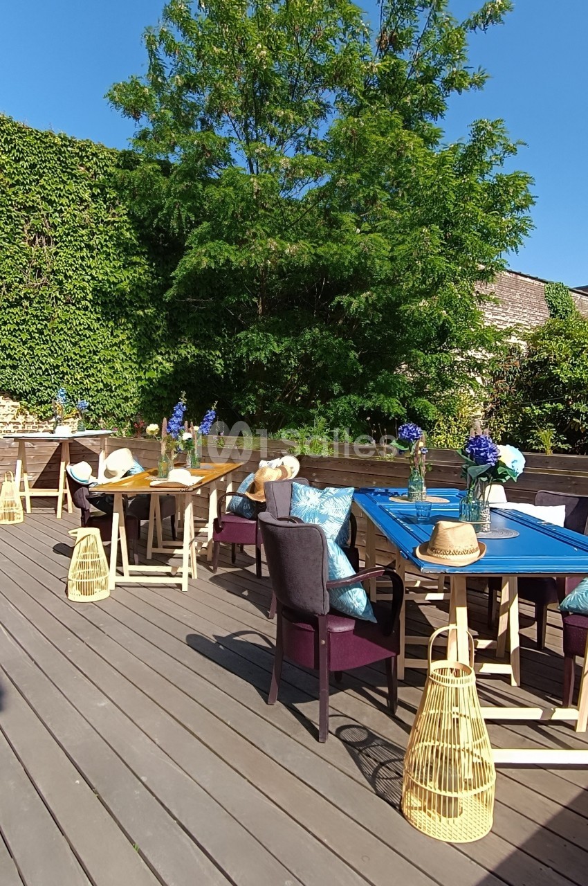 Terrasse en bois aménagée avec des tables, chaises et décorations, entourée de verdure sous un ciel bleu.
