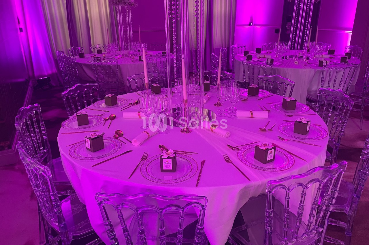 Table ronde décorée pour un événement, avec nappes blanches, couverts dorés, bougies et éclairage violet.