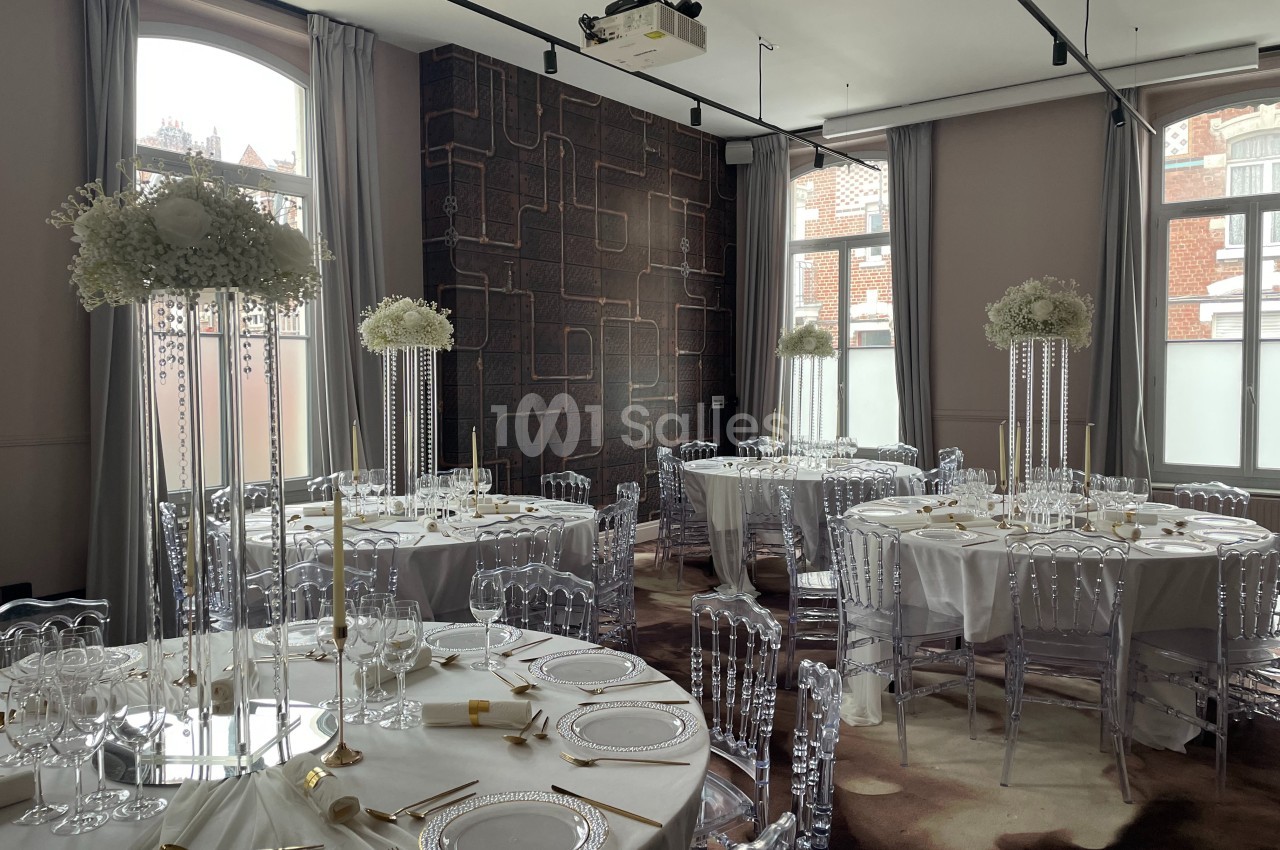 Salle de réception élégamment décorée avec des tables rondes, nappes blanches, chaises transparentes et centres de table…