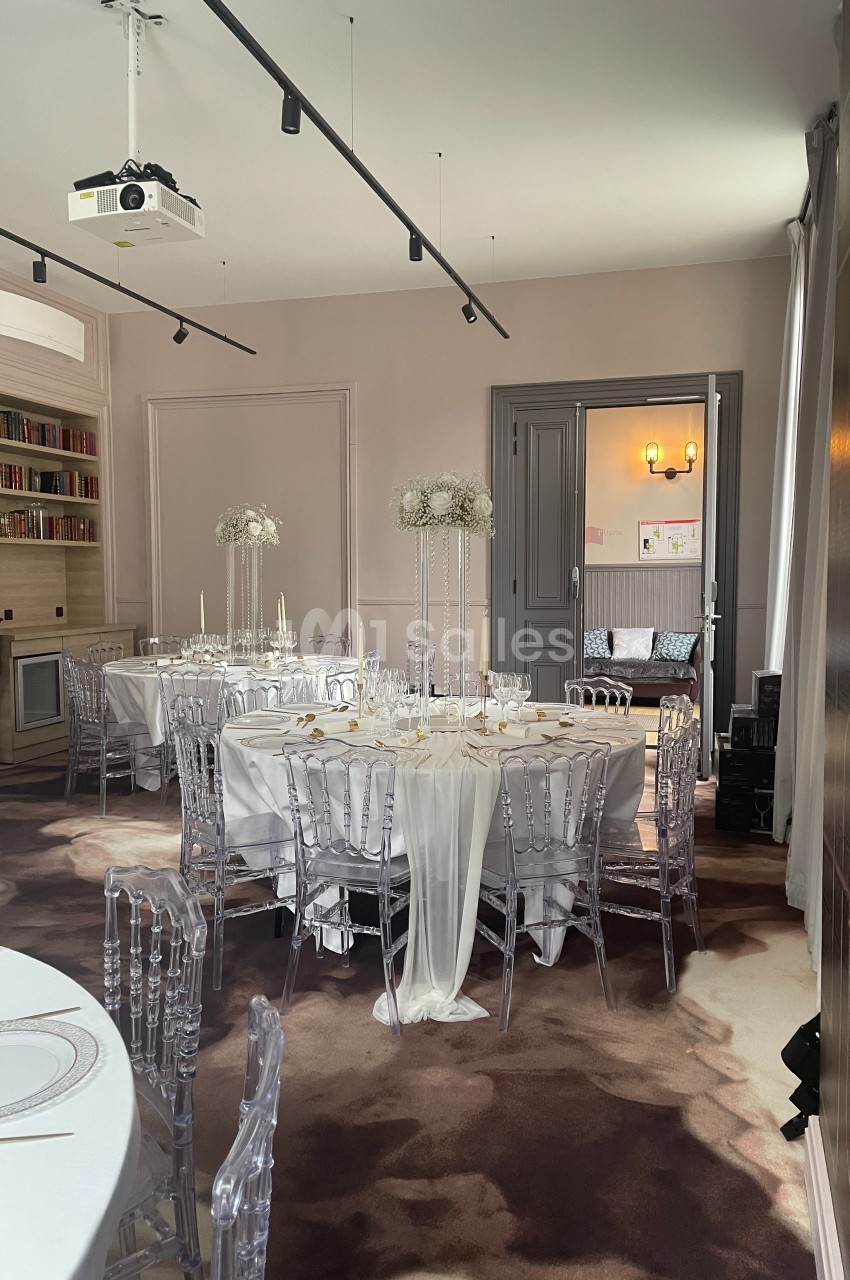 Salle élégante décorée pour un événement, avec tables rondes, nappes blanches, chaises transparentes et centres de table…