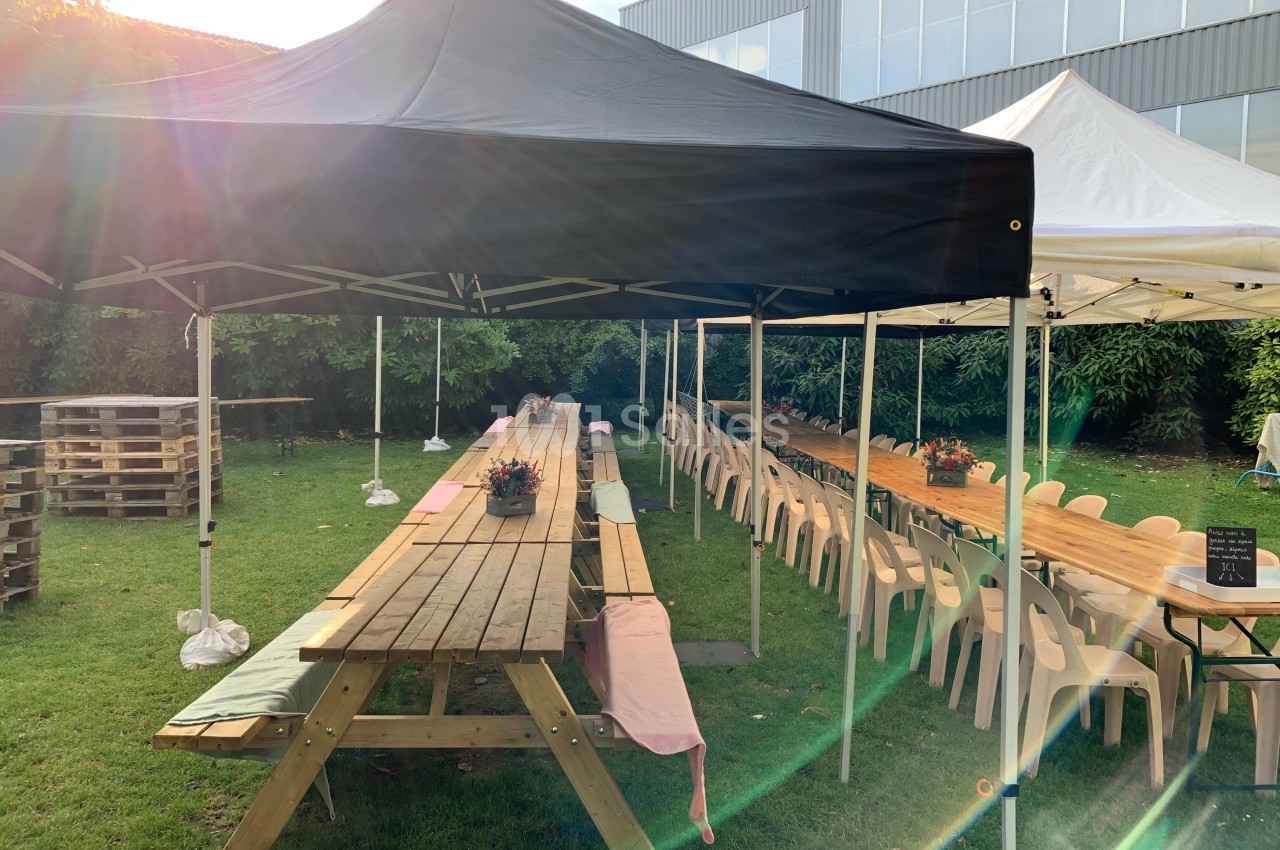 Tables en bois alignées sous des tentes noires et blanches dans un jardin, entourées de chaises et de verdure.