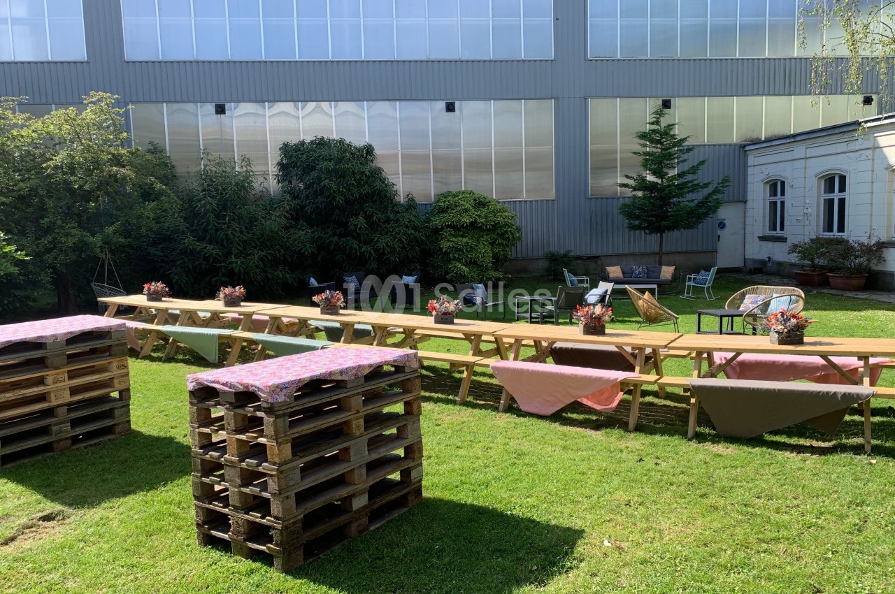Tables en bois décorées de nappes et de bouquets, disposées sur une pelouse devant un bâtiment industriel.