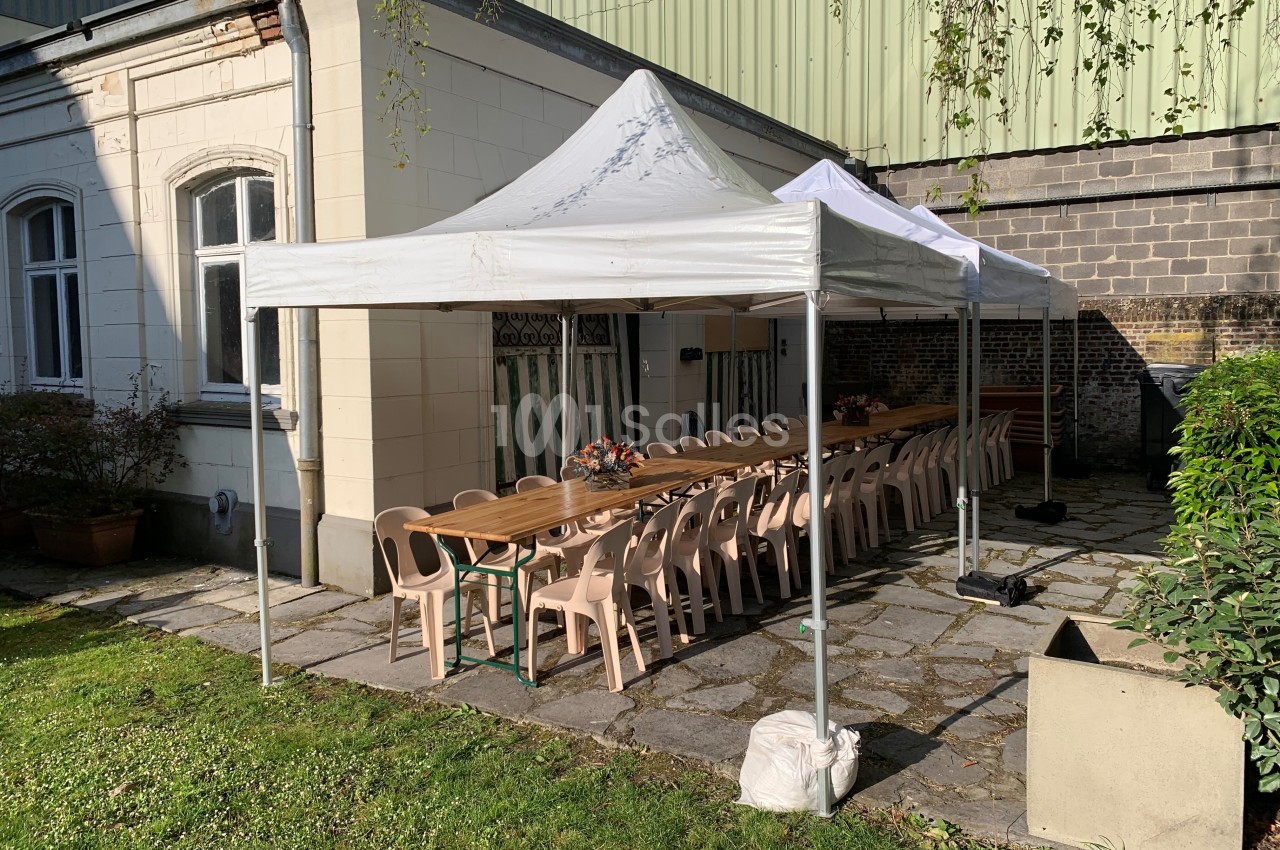 Deux tentes blanches abritent une longue table avec des chaises en plastique beige, installées dans une cour pavée.