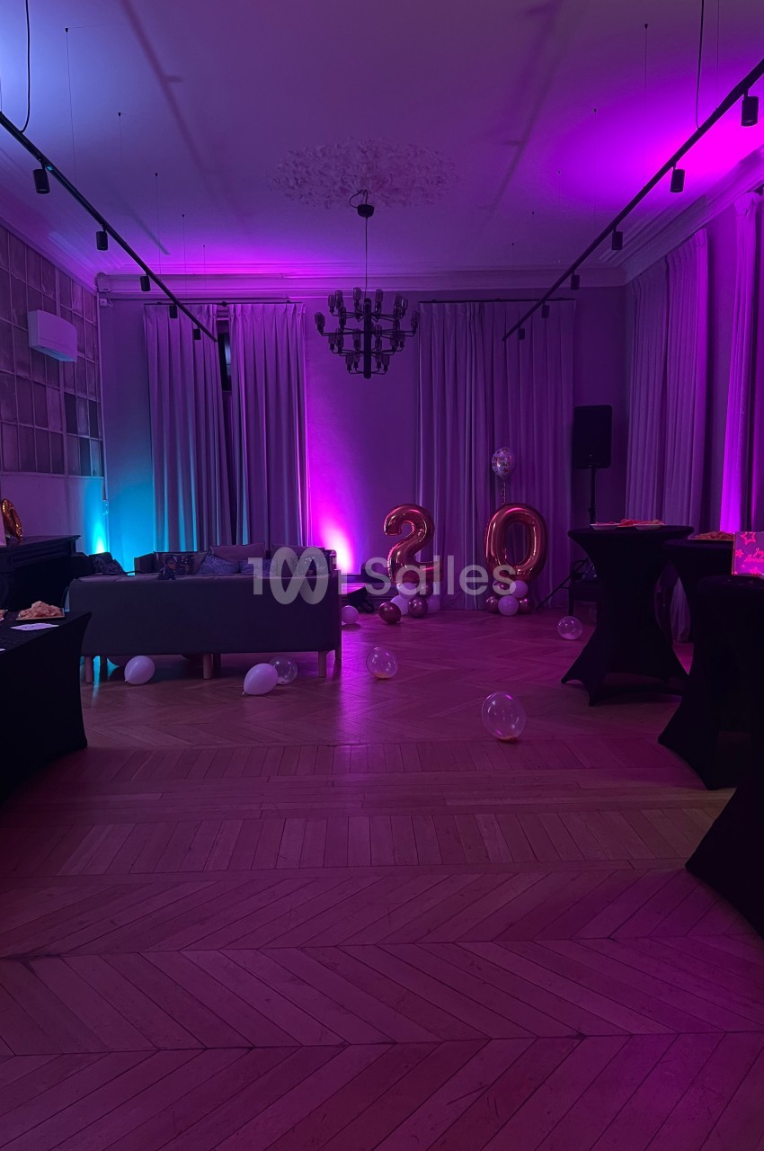 Salle décorée pour une fête avec éclairage violet, ballons ’20’, tables hautes et canapé en arrière-plan.