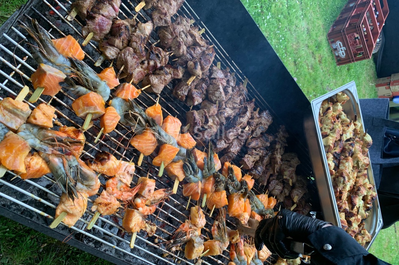 Brochettes de crevettes, saumon et viande grillant sur un barbecue en plein air, avec un plateau de viande à côté.